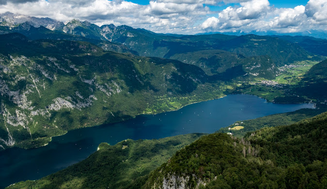 "Mountainous landscape featuring a winding river or lake. The image showcases lush green hills, rugged mountains, and a bright blue sky with scattered clouds. A white cursor is visible on the screen, pointing to an area in the landscape."